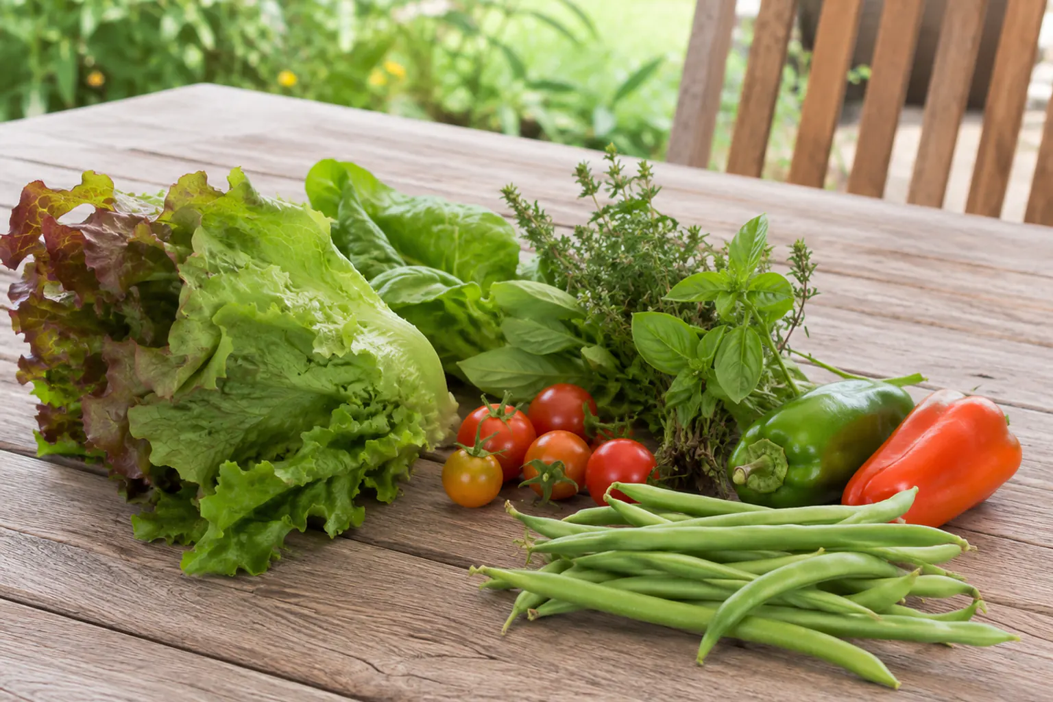 A small freshly picked selection of high-value crops on a wooden table: leaf lettuces, herbs, cherry tomatoes, green beans, and sweet peppers.