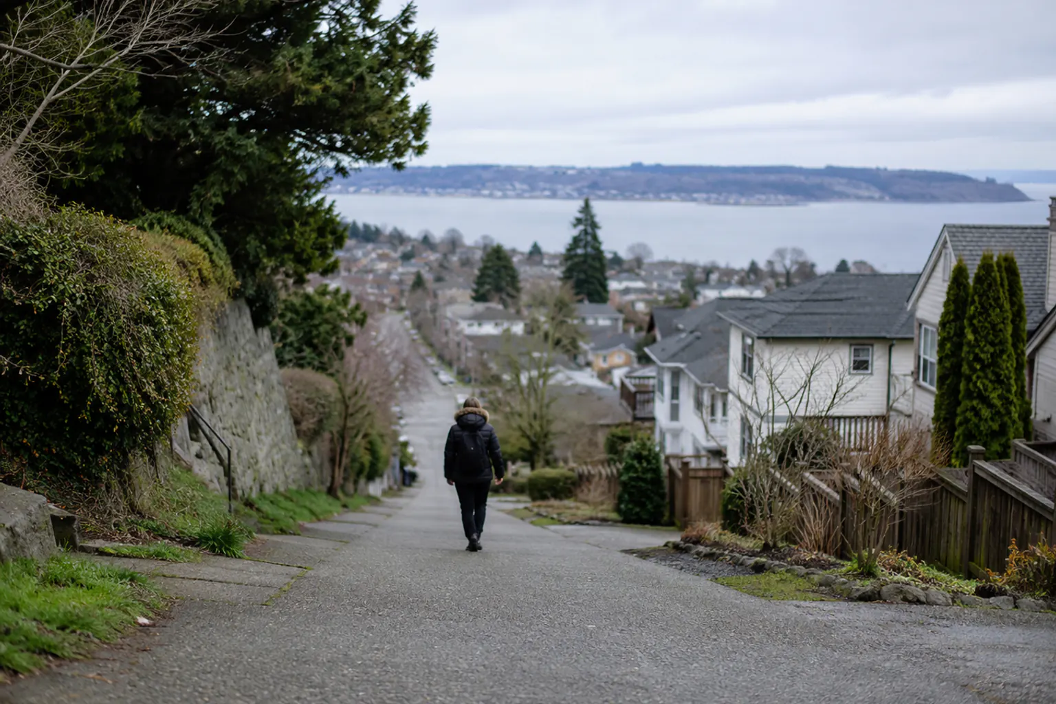 A sloping residential street leading uphill and away from a low area toward higher ground, a figure walking up seen from behind on an overcast day.