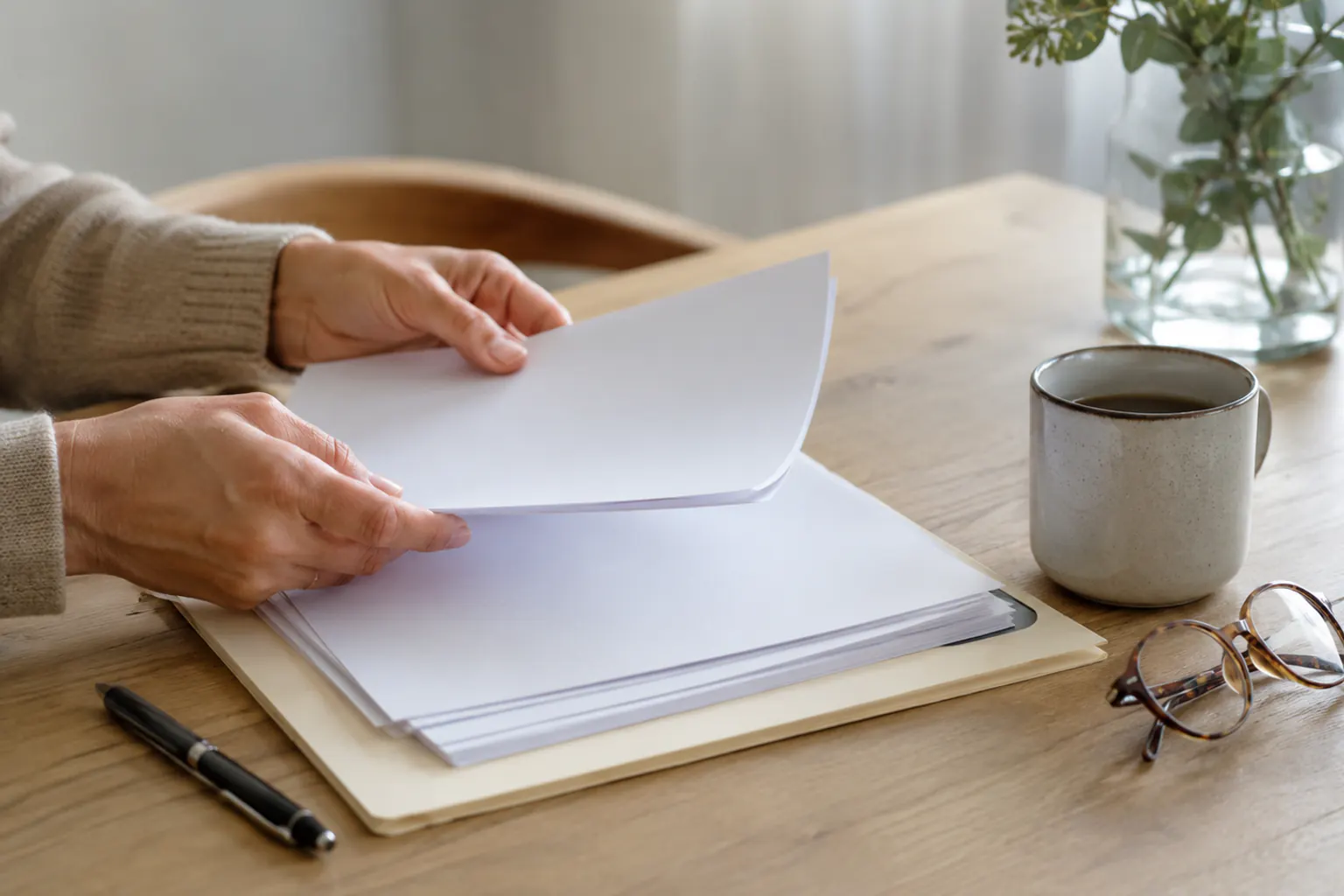 Hands leafing through blank pages in a folder on a wood table beside a mug, reading glasses, and a vase of greenery.