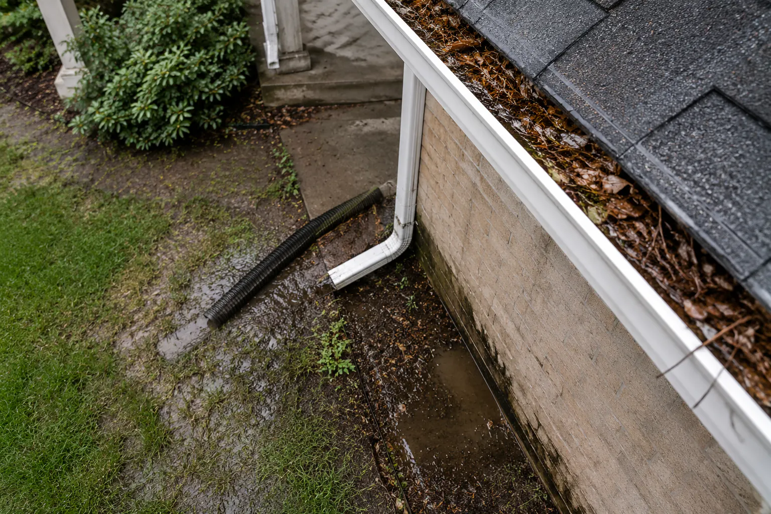 A downspout discharging beside a house foundation with visible soil erosion, photographed from ground level.