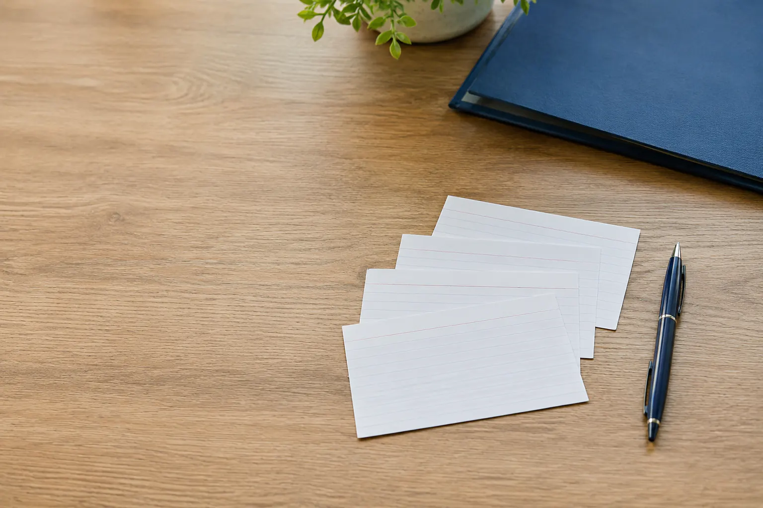 Four blank index cards fanned out on a table beside a pen, with a folder and a small plant nearby.