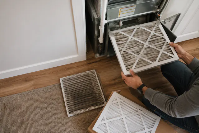 Person holding a used HVAC air filter beside a return grille.