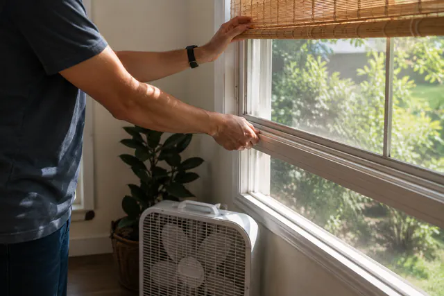 Person lowering a window shade above an indoor fan.