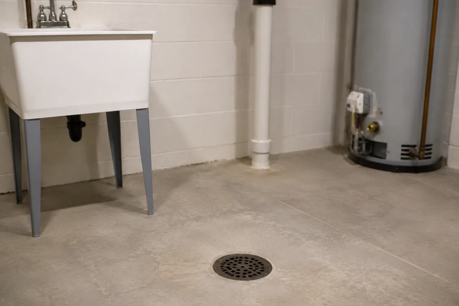A clean, dry basement floor drain on bare concrete near a utility sink and water heater.