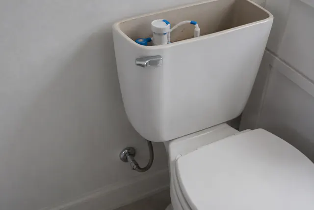 A closed bucket-style emergency toilet with a lid and a sealed bag supply in a plain bathroom corner, no waste visible.
