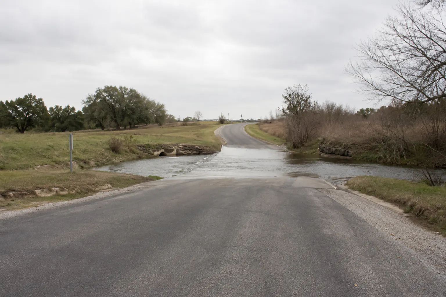 A rural low-water road crossing where shallow water covers the road ahead, seen from a stopped distance well back on an overcast day.